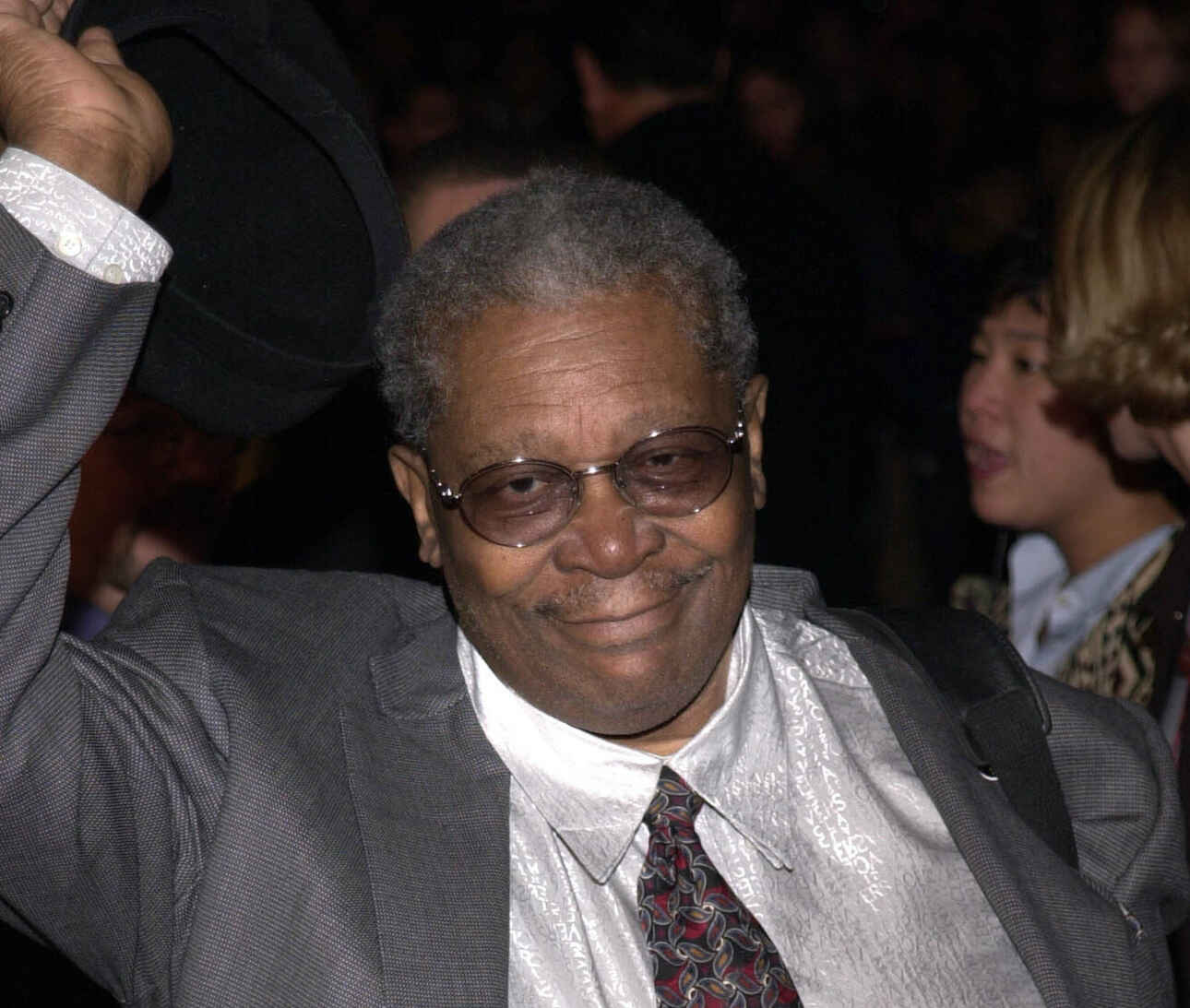 BB King, wearing a grey suit and a brown tie, is waving his hand.