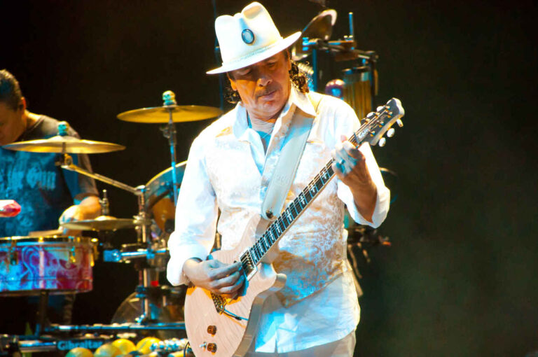 Carlos Santana live on stage wearing a white suit and white hat.