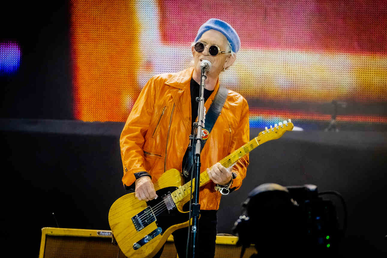 Keith Richards singing, wearing sun glasses and a blue bandana while playing his yellow Telecaster with black pickguard during a Rolling Stones concert.