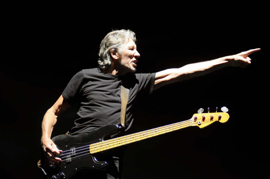 Roger Waters playing a black Fender Precision bass live on stage, while pointing with his left hand.